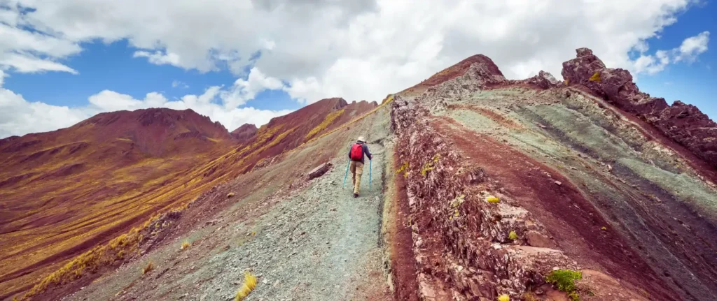 Person hiking along the colorful, jagged ridge of Pallay Punchu, offering a remote and scenic trail compared to Vinicunca in the Rainbow Mountain debat